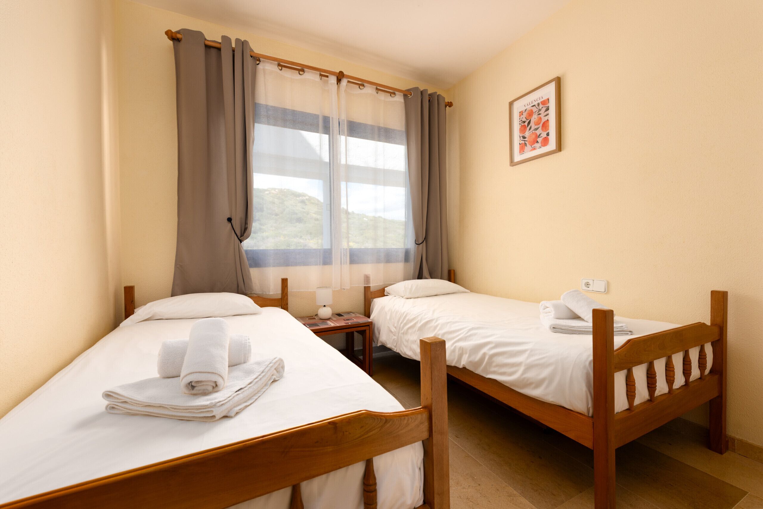 A room with two wooden beds, a window with curtains, a wall-mounted light, and a framed picture.