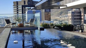 Una piscina en la azotea con sillones de descanso, vista panorámica de la ciudad y un edificio con el logotipo 'absa'.