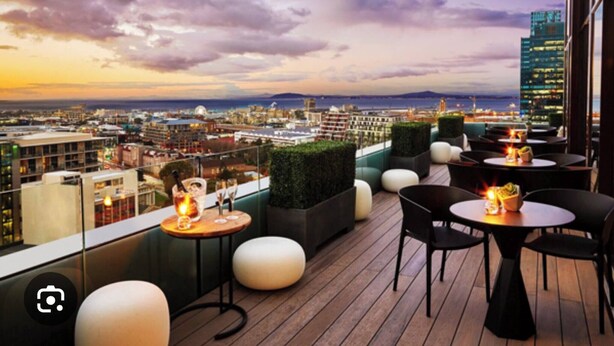 Terraza en la azotea con mesas y sillas, con vista a un paisaje urbano al atardecer.