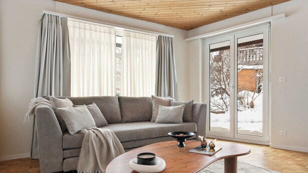 Ein Wohnzimmer mit grauem Sofa, einem Holztisch und einem großen Fenster mit Vorhängen.