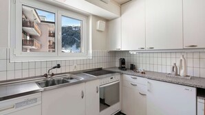 Eine moderne Küche mit weißen Schränken, einem eingebauten Backofen, einem Geschirrspüler und einem großen Fenster mit Blick auf einen Balkon und Bäume.