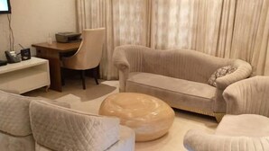 Un soggiorno con un divano color beige, un pouf rotondo e una poltrona. Si nota una scrivania con una sedia e un televisore fissato alla parete.