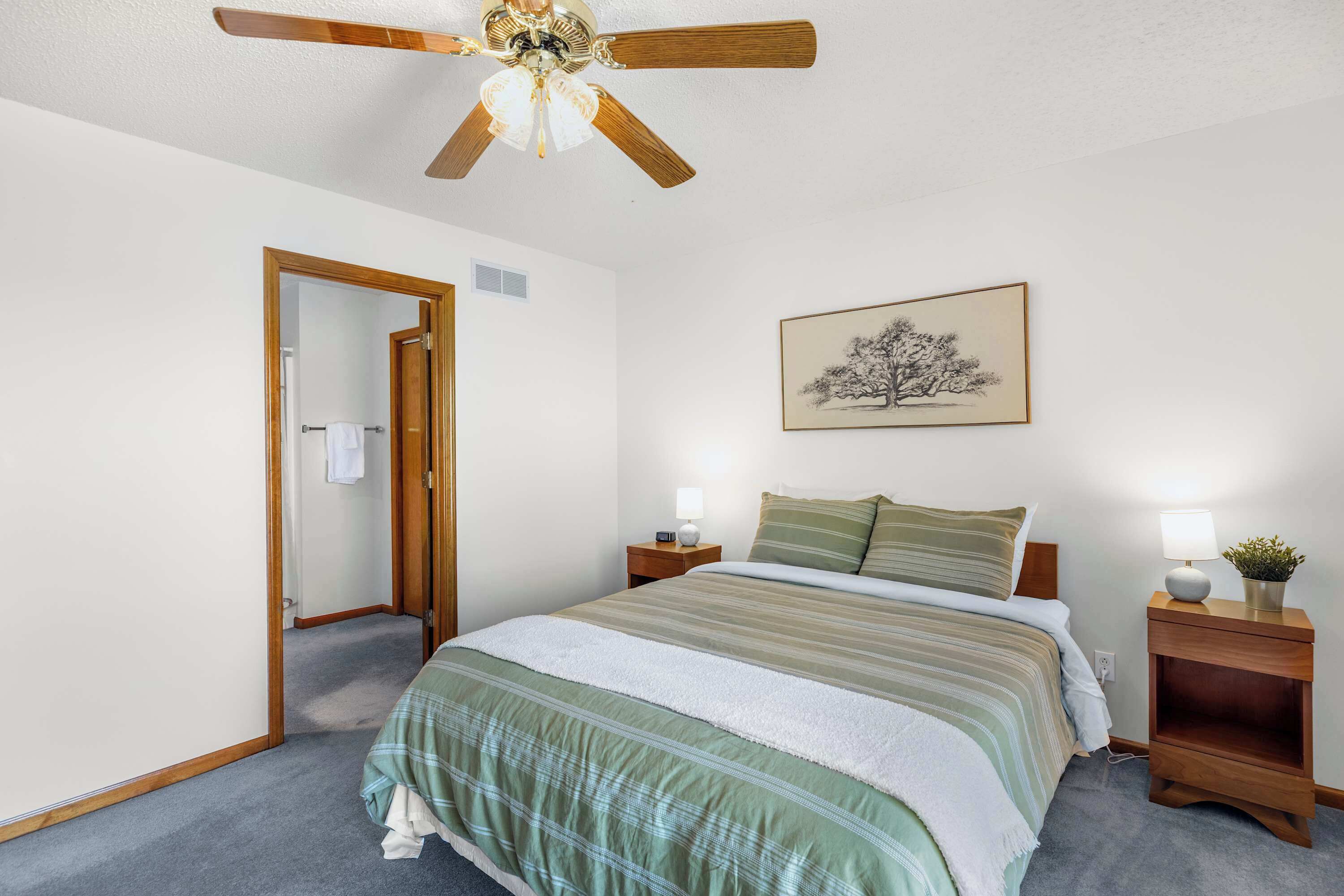 A bedroom with a bed, a ceiling fan, a nightstand with a lamp, a framed picture on the wall, and a door leading to another room.