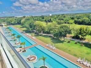 A long swimming pool with lounge chairs and palm trees, surrounded by lush greenery and a distant building. - Luxe Condo with/Rooftop @ Star (Bavaro)