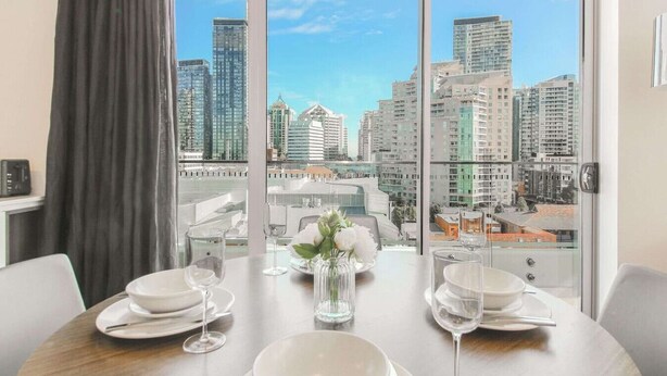 Un comedor con una mesa lista para dos personas, un florero con flores blancas y vista a la ciudad a través de amplias ventanas.