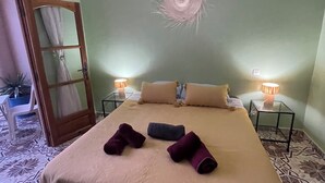 A neatly made bed with a beige bedspread, two rolled towels, and two pillows. There are two bedside tables with lamps, a mirror, and a door leading to another room.