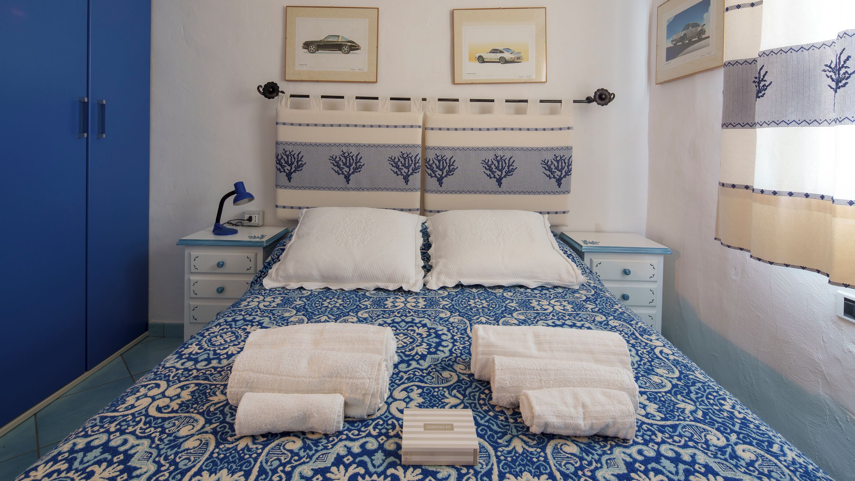 A neatly made bed with a patterned bedspread, two pillows, and four folded towels. There are two bedside tables with lamps, a framed picture on the wall, and a blue wardrobe.