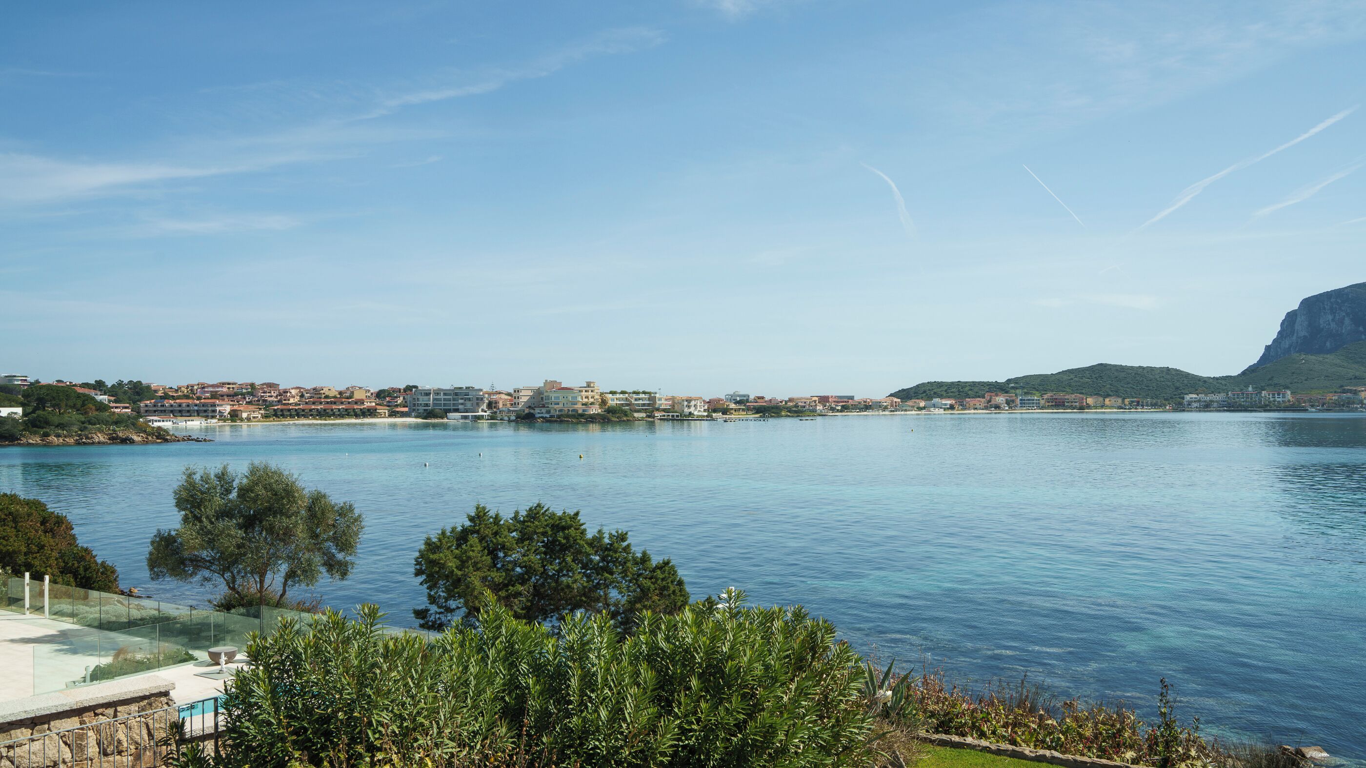 A coastal town with buildings along the water, a clear sky, and a mountain in the background.