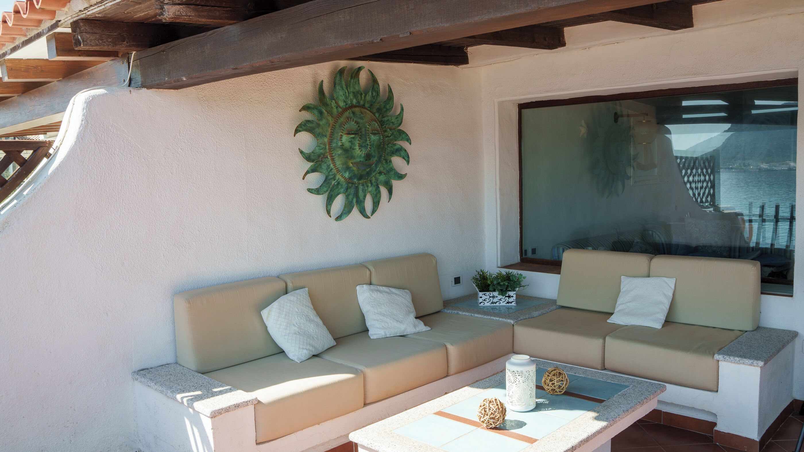 A sunlit outdoor seating area with two beige sofas, a small table with decorative items, and a large window offering a view of a body of water and mountains.