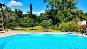 A rectangular swimming pool with a wooden deck surrounding it, set in a lush garden with trees and a clear blue sky. - House in the heart of Provence (Flayosc)