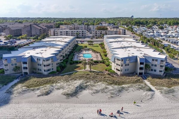 Un hotel frente a la playa con piscina central, varios balcones y una zona de arena.