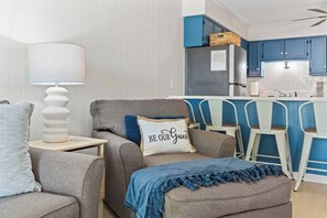 Una sala de estar con un sofá, una mesita auxiliar y una lámpara. Hay una zona de cocina con armarios azules y sillas blancas.