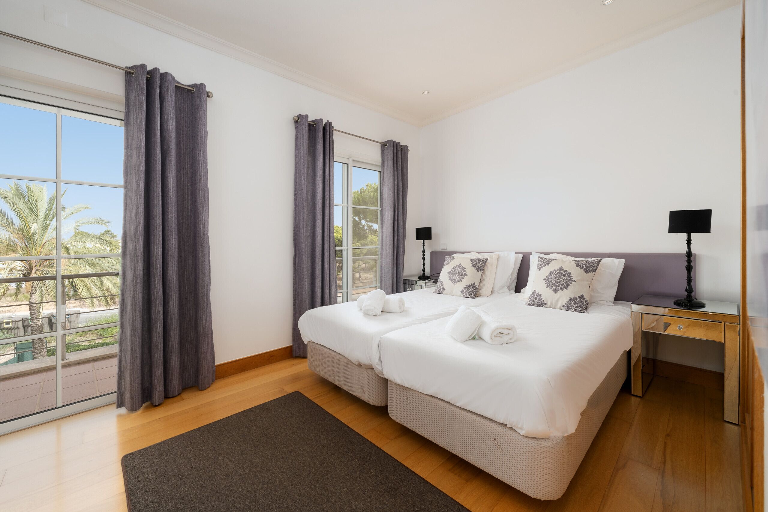 A double bed with white linens and decorative pillows, a bedside table with a lamp, and a view of palm trees through large windows.