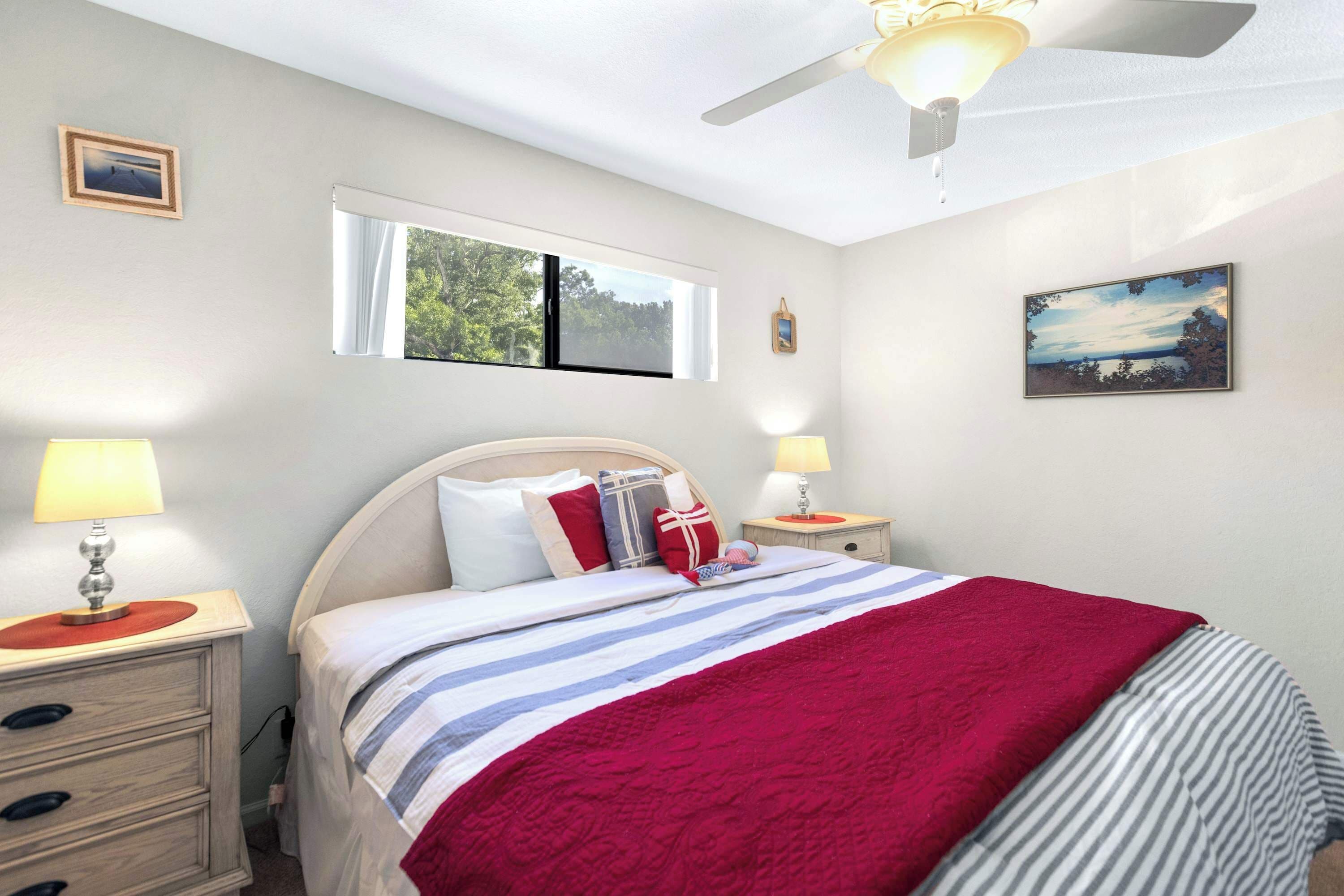 Une chambre à coucher avec un lit, deux tables de chevet avec des lampes, un ventilateur de plafond, une fenêtre donnant sur des arbres et un tableau accroché au mur.