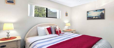 Une chambre à coucher avec un lit, deux tables de chevet avec des lampes, un ventilateur de plafond, une fenêtre donnant sur des arbres et un tableau accroché au mur.