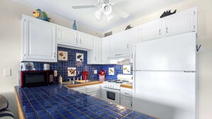 Una cocina con armarios blancos, salpicadero de azulejos azules, microondas, estufa y refrigerador.