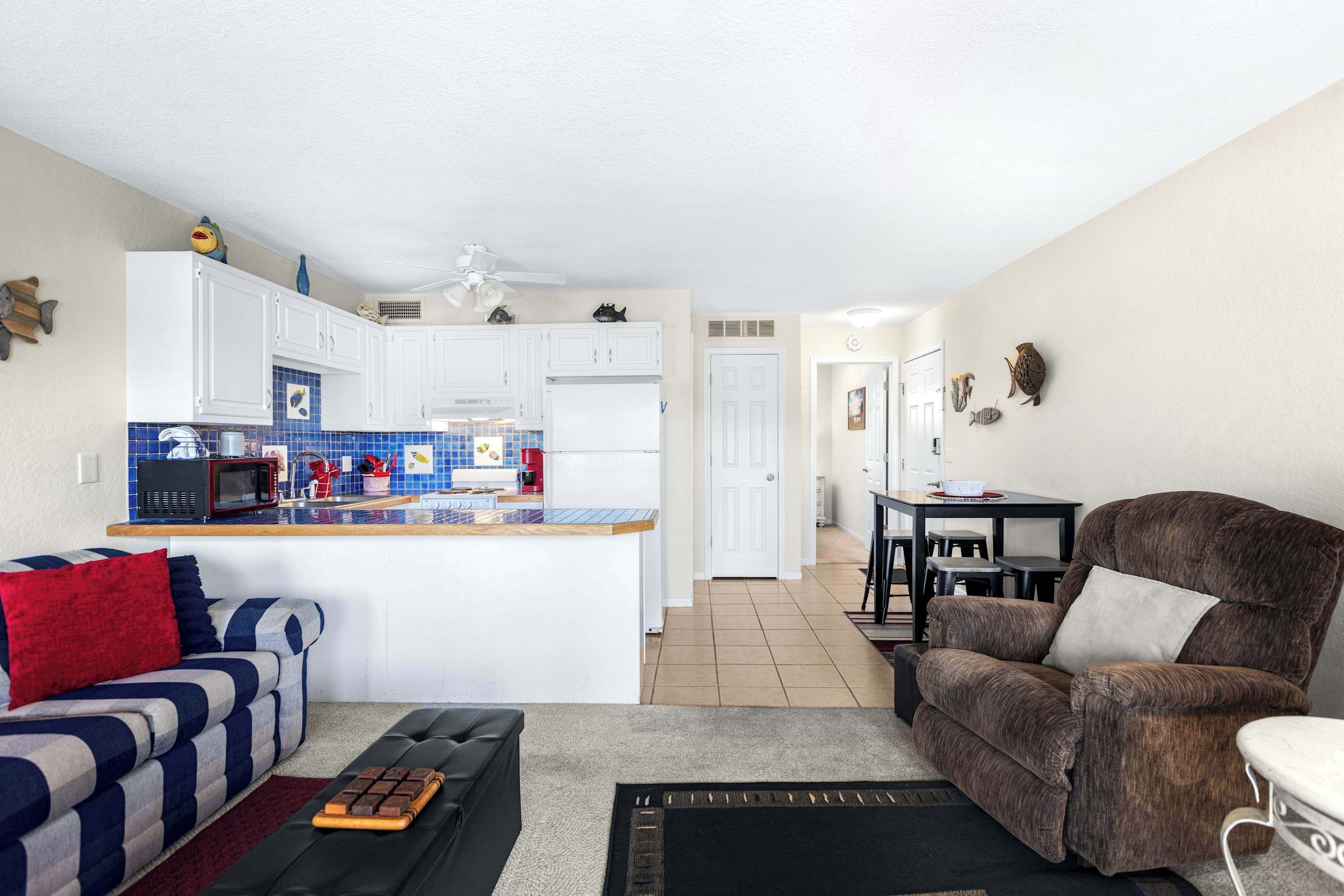 A living room with a sofa, a brown armchair, a coffee table, and a kitchen area with white cabinets and a blue backsplash.