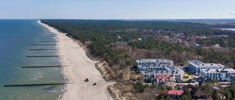 An aerial view of a coastal area with a sandy beach, a row of buildings with multiple stories, and a forested area in the background.