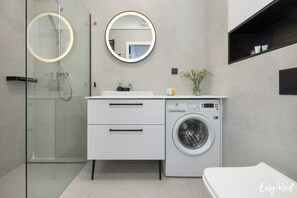 Une salle de bain moderne avec une douche à parois de verre, un miroir rond, un lave-linge encastré et un meuble-lavabo blanc doté de tiroirs.
