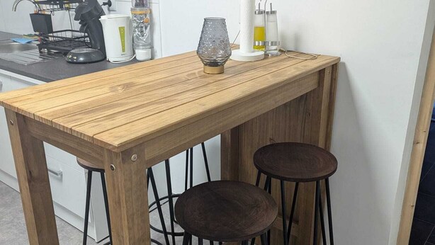 A wooden table with two stools, a kitchen area with a sink and utensils, and a white wall with an electrical outlet. - Apartamento a 5 minutos de Luxemburgo fully equipped kitchen, lovely terrace (Audun-le-Tiche)