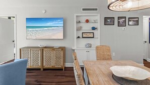 Un coin repas avec une table en bois, des chaises en osier, un grand tableau sur le thème de la plage et un luminaire suspendu en osier.