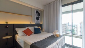 A modern hotel room with a large bed, a nightstand, a small table, and a window with a city view. - Modern Retreat Near Allianz Parque Sp-p1604a (São Paulo)
