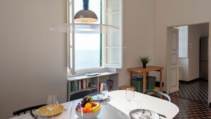 A dining area with a white table, clear glass pendant lights, a bowl of fruit, and a view of the sea through an open window.