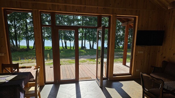 Una cabaña de madera con un gran ventanal corredera que da a una terraza exterior. En el interior, hay una mesa de comedor con sillas y un televisor de pantalla plana montado en la pared.