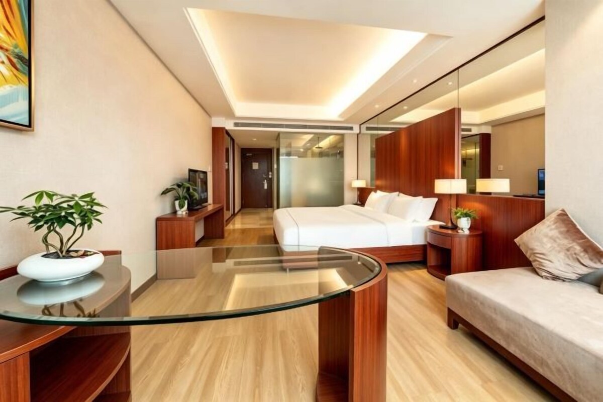 A modern hotel room with a glass coffee table, a bed, a sofa, a desk, and a plant.