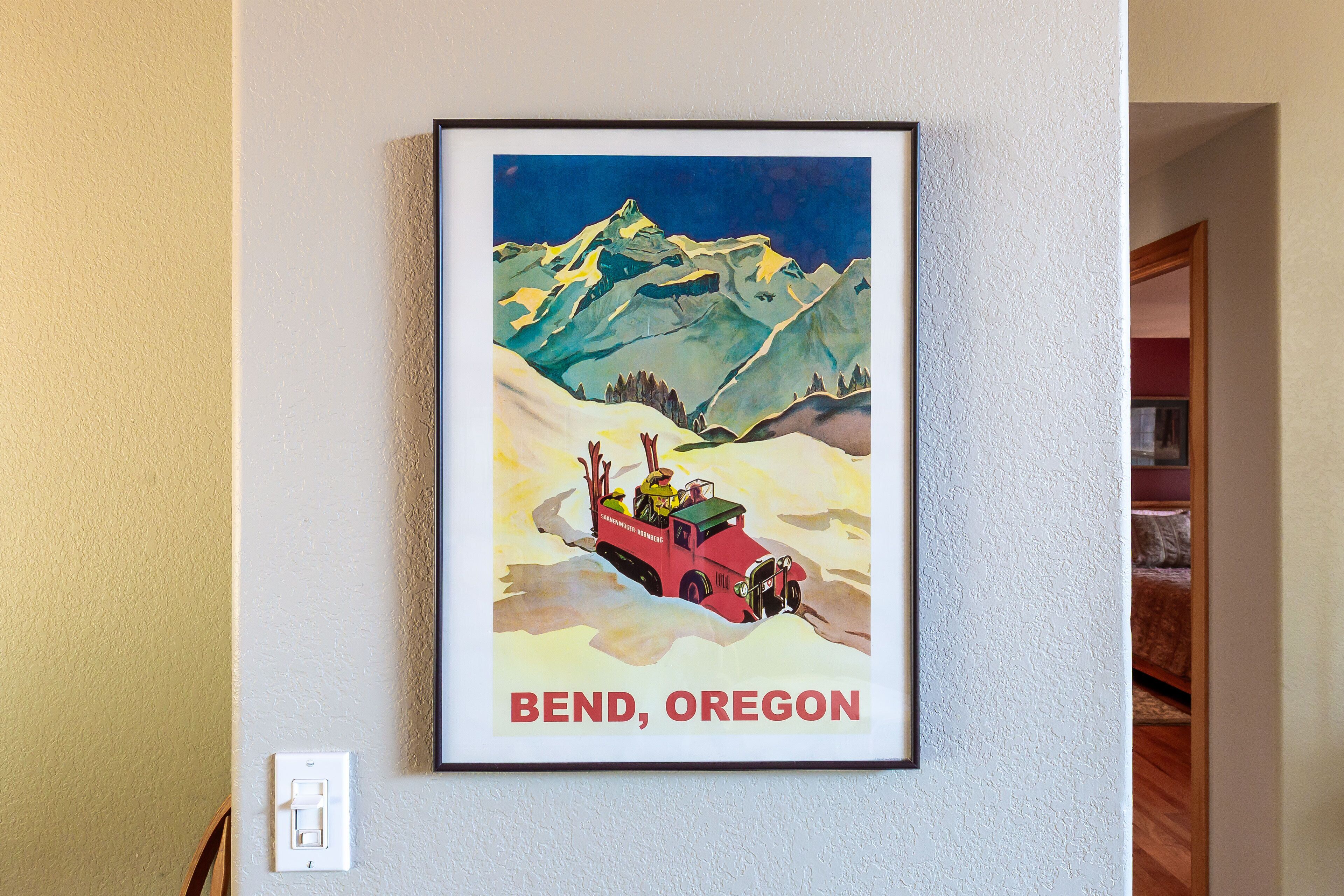 Um cartaz emoldurado com uma paisagem de montanha, um veículo vermelho e a inscrição 'Bend, Oregon' na parede.