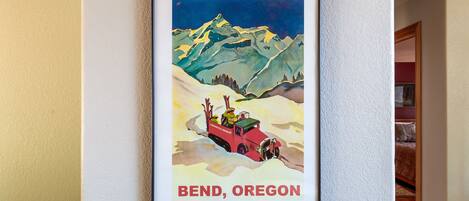Um cartaz emoldurado com uma paisagem de montanha, um veículo vermelho e a inscrição 'Bend, Oregon' na parede.