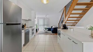 A modern kitchen with stainless steel appliances, a dining area with a dark-colored sofa, and a staircase with wooden railings. - Central Redcliffe Coastal Home Walk to Beach (Redcliffe)