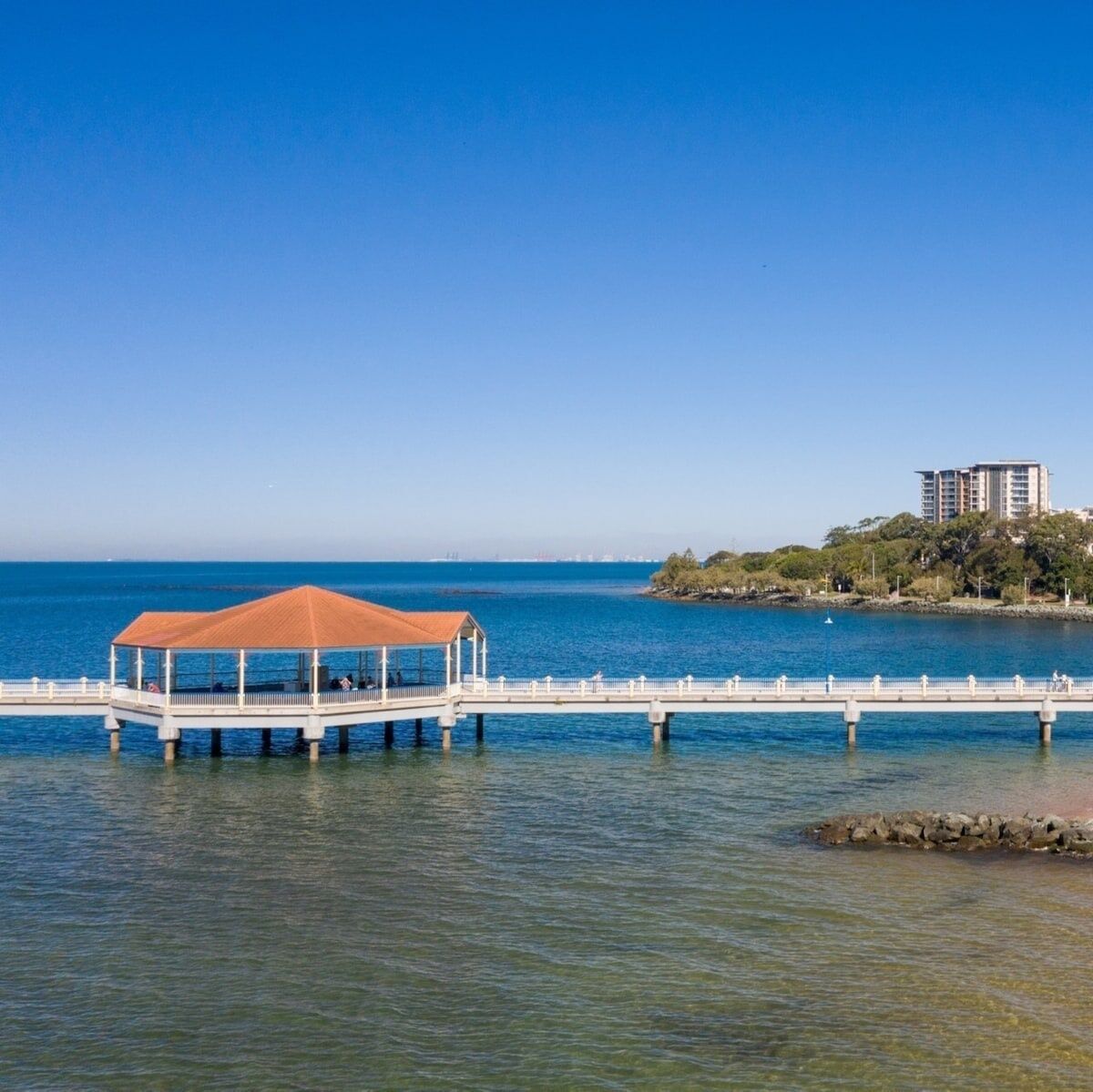 Un muelle con una zona de estar cubierta que se extiende sobre un cuerpo de agua, con una vista distante de un conjunto de edificios.