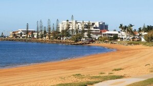 A coastal beach with a sandy shore, a rocky breakwater, and residential buildings in the background. - Central Redcliffe Coastal Home Walk to Beach (Redcliffe)