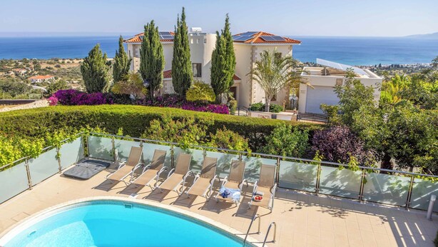 A poolside area with lounge chairs, a clear view of the sea, and a building with solar panels. - Villa Prengos 6 (Neo Chorio)