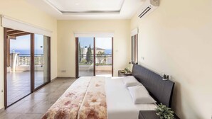 A bedroom with a bed, a nightstand, a potted plant, and a view of the sea through large sliding doors. - Villa Prengos 6 (Neo Chorio)