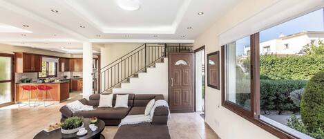 A spacious living area with a sofa, ottoman, and coffee table, a kitchen in the background, and a staircase leading to an upper floor.