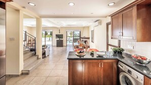 A modern kitchen with wooden cabinets, a built-in washing machine, and a dining area in the background. - Villa Prengos 6 (Neo Chorio)