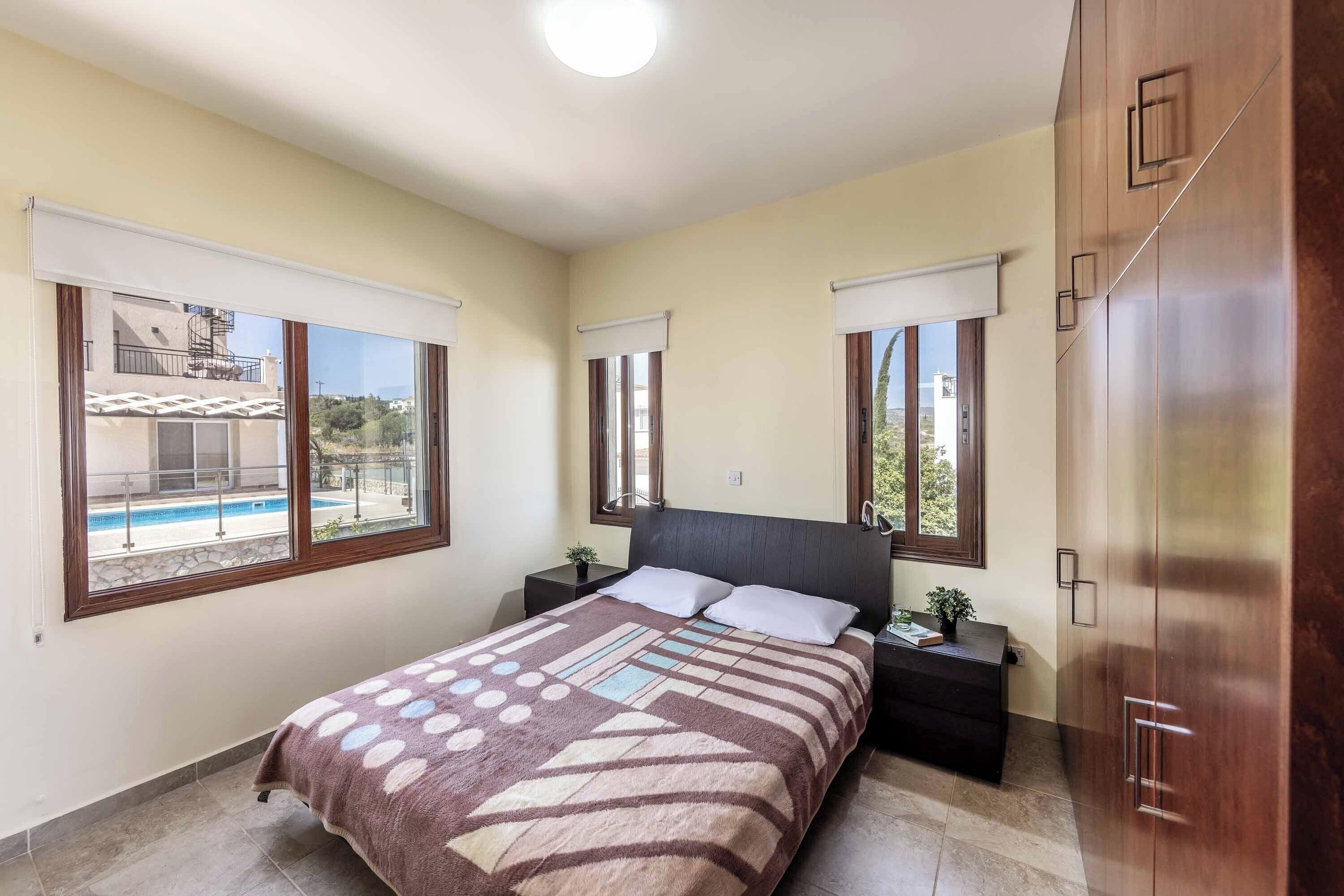 Une chambre à coucher comprenant un lit, deux tables de chevet, une commode, une armoire et de grandes fenêtres équipées de stores.