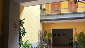 Un patio interior amplio con suelo de baldosas, rodeado de plantas en macetas y un balcón con persianas de madera.