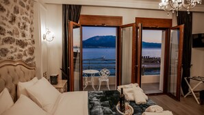 Habitación de hotel con una cama, una mesita con una botella y vasos, y vistas a un lago y montañas a través de puertas abiertas.