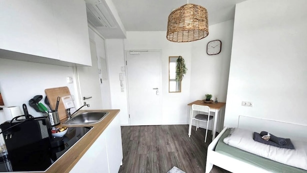 Een compacte keuken met een zwart inductiefornuis, een houten aanrechtblad en een klein zite hoekje met een witte tafel en stoel. Een geweven hanglamp bevindt zich boven het zithoekje, en er ligt een grijs vloerkleed op de vloer.