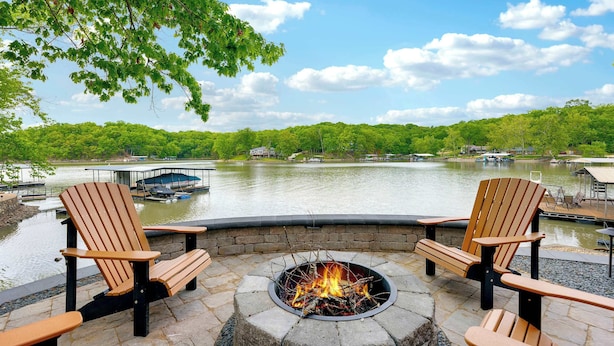 A lakeside patio with Adirondack chairs, a stone fire pit, and a view of the lake and boats. - Trails End Hideaway Waterfront (Sunrise Beach)