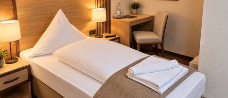 A hotel room with a neatly made bed, a bedside lamp, a small desk with a chair, and a potted plant.
