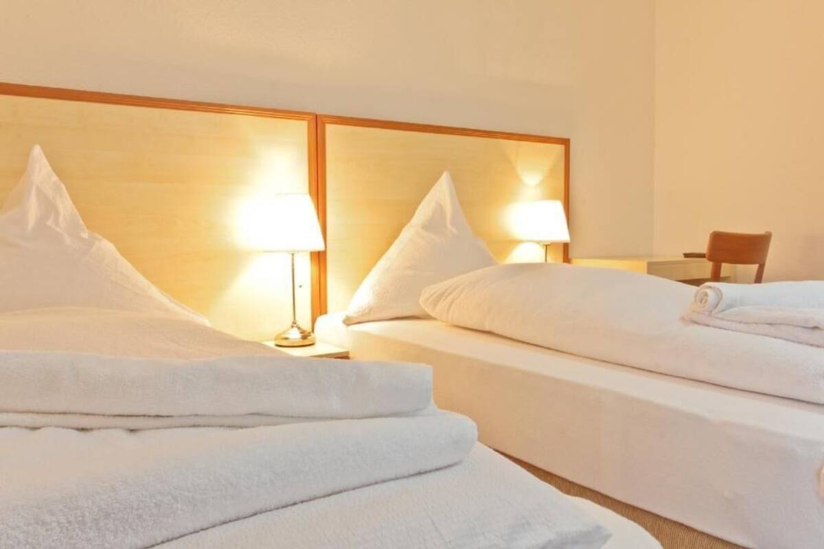 Habitación de hotel con dos camas, cada una con una almohada y una toalla doblada. Hay dos lámparas de noche y una silla en la esquina.