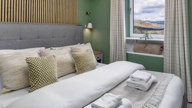 A neatly made bed with pillows and folded towels, a bedside lamp, and a view of the outdoors through a window with curtains. - Ochill View Apartment by Stirling Train Station (Stirling)