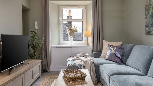 A living room with a TV, sofa, and window with curtains. - Central Cathedral Apartment (Dunblane)
