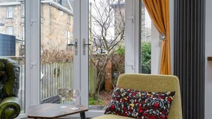 A sun lounger with a patterned pillow, a small table with a glass, and a view of a garden through a glass door.