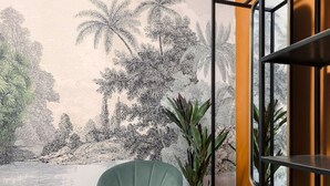 Una habitación con un sillón verde, una estantería metálica negra y un mural de paisaje tropical.