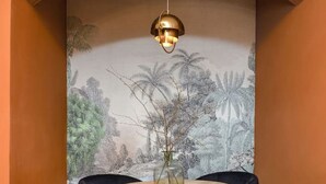 Un comedor con una mesa redonda de madera, dos sillas tapizadas de color negro, un jarrón con flores secas y un mural de follaje tropical en la pared.